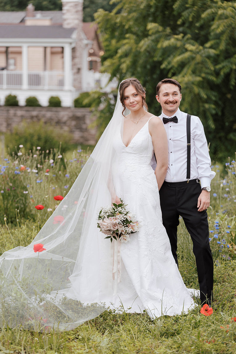 Bride and groom standing in a field, smiling, with wedding attire and flowers.