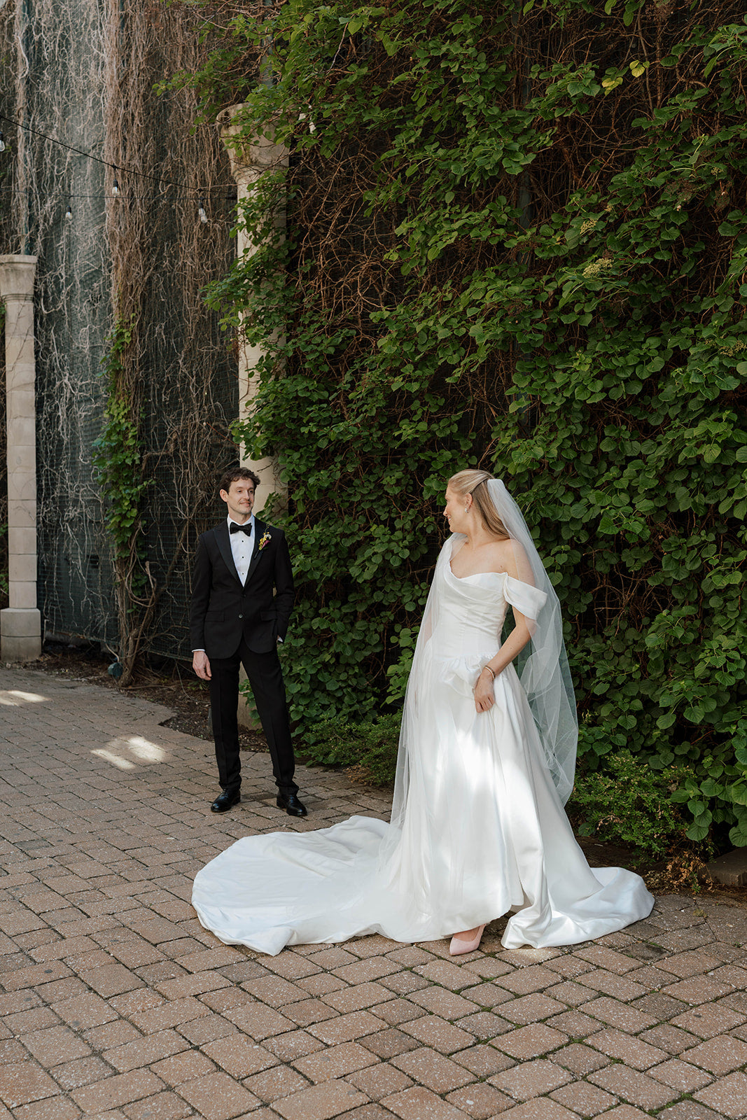 Bride in white dress and groom in tuxedo near greenery.