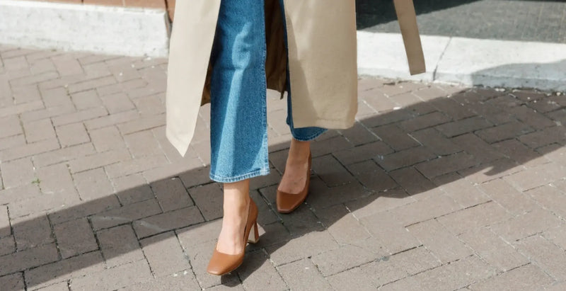 Woman wearing cropped jeans, a beige coat, and brown block-heel pumps from ALLY Shoes while walking on a city sidewalk.